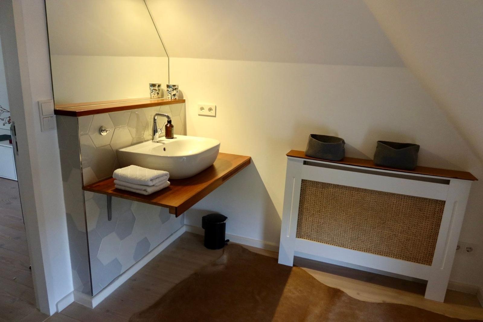 Waschbecken mit Spiegel und Holzregal. Bodenbelag aus Holz und Fell. Wandheizung mit Körbchen.