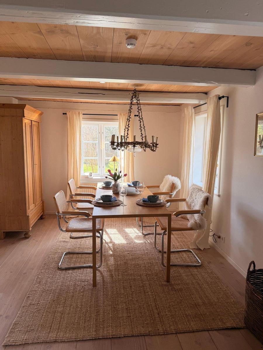 Esszimmer mit Holztisch, Stühlen und Kronleuchter. Holzdecke und Fenster mit Vorhängen.