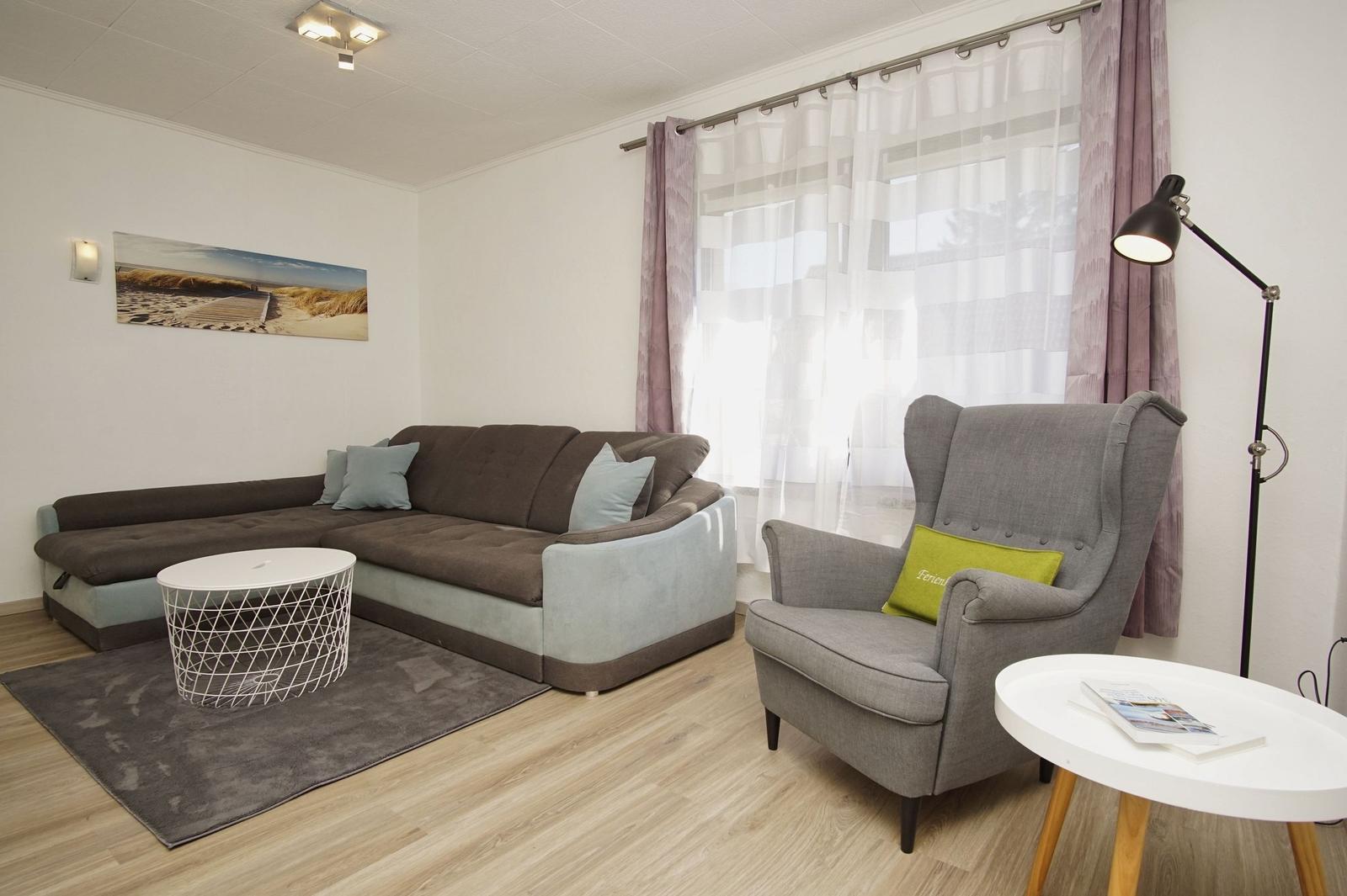 Wohnzimmer mit Ecksofa, Sessel, Tischen und Vorhängen vor Fenster.
