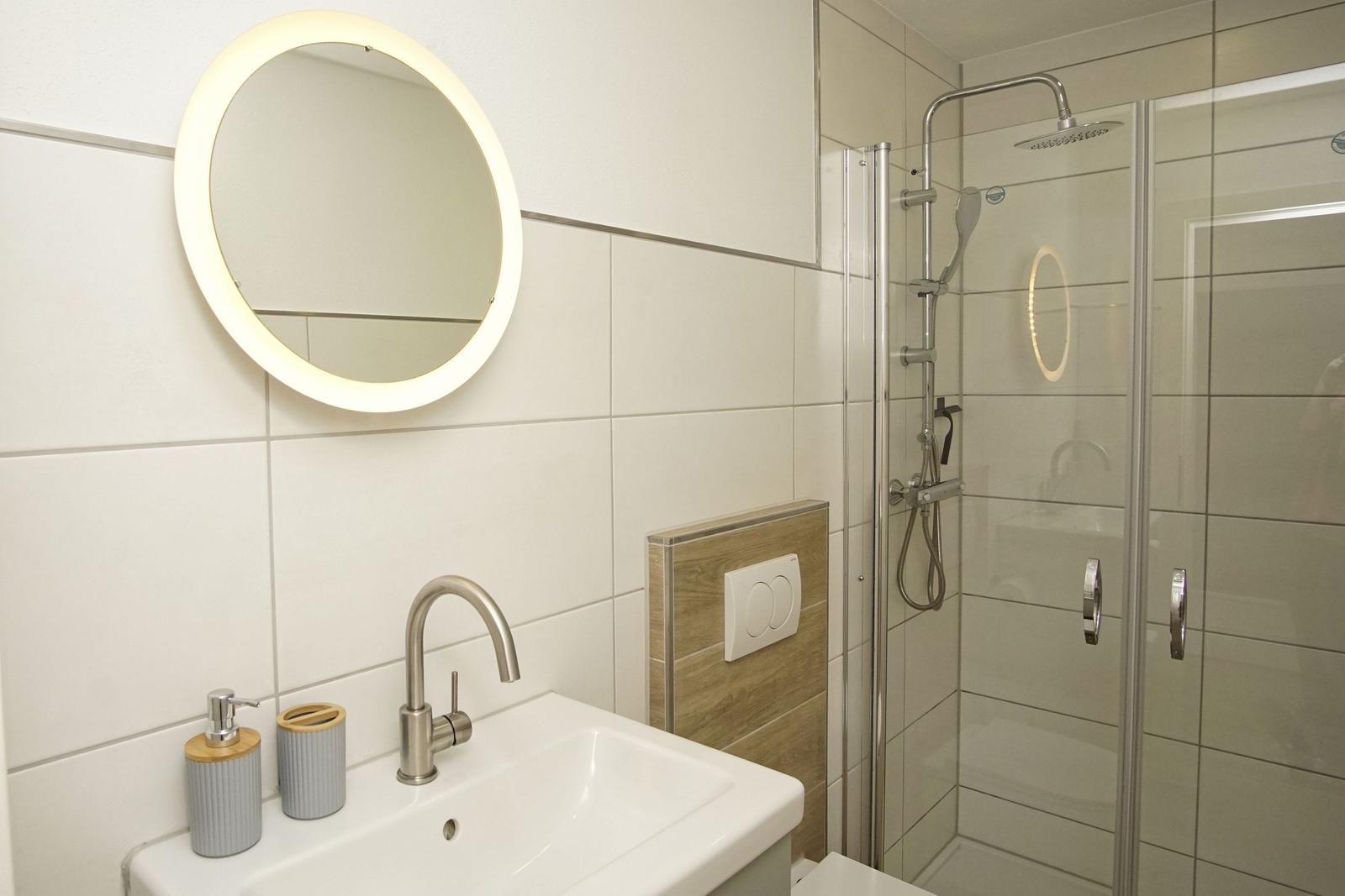 Badezimmer mit Dusche, Waschbecken und runden Spiegel mit LED-Beleuchtung.