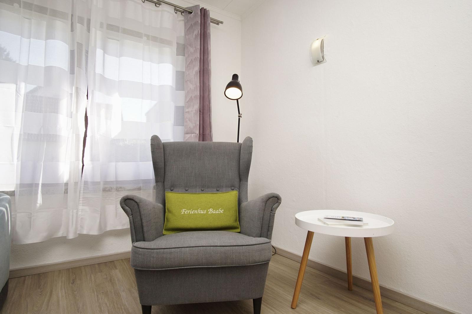 Graue Sessel mit grünem Kissen, kleiner runder Tisch und Stehlampe neben Fenster mit Vorhängen.