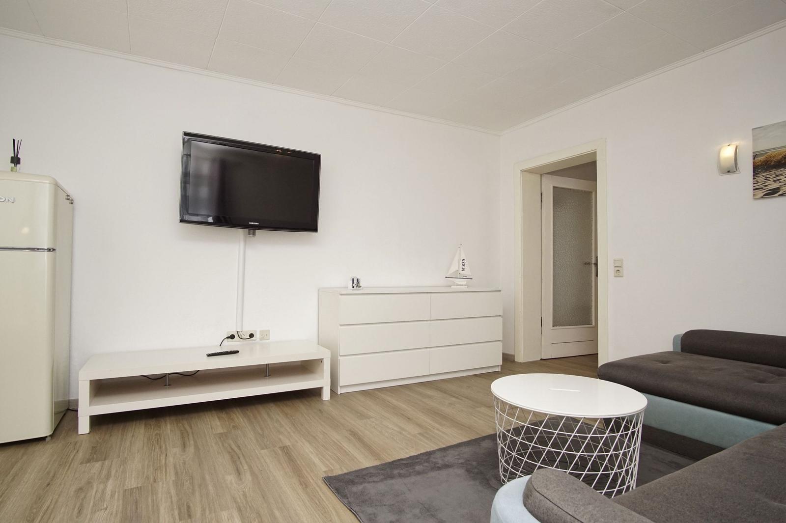 Wohnzimmer mit Sofa, Fernseher, Sideboard und Kühlschrank.