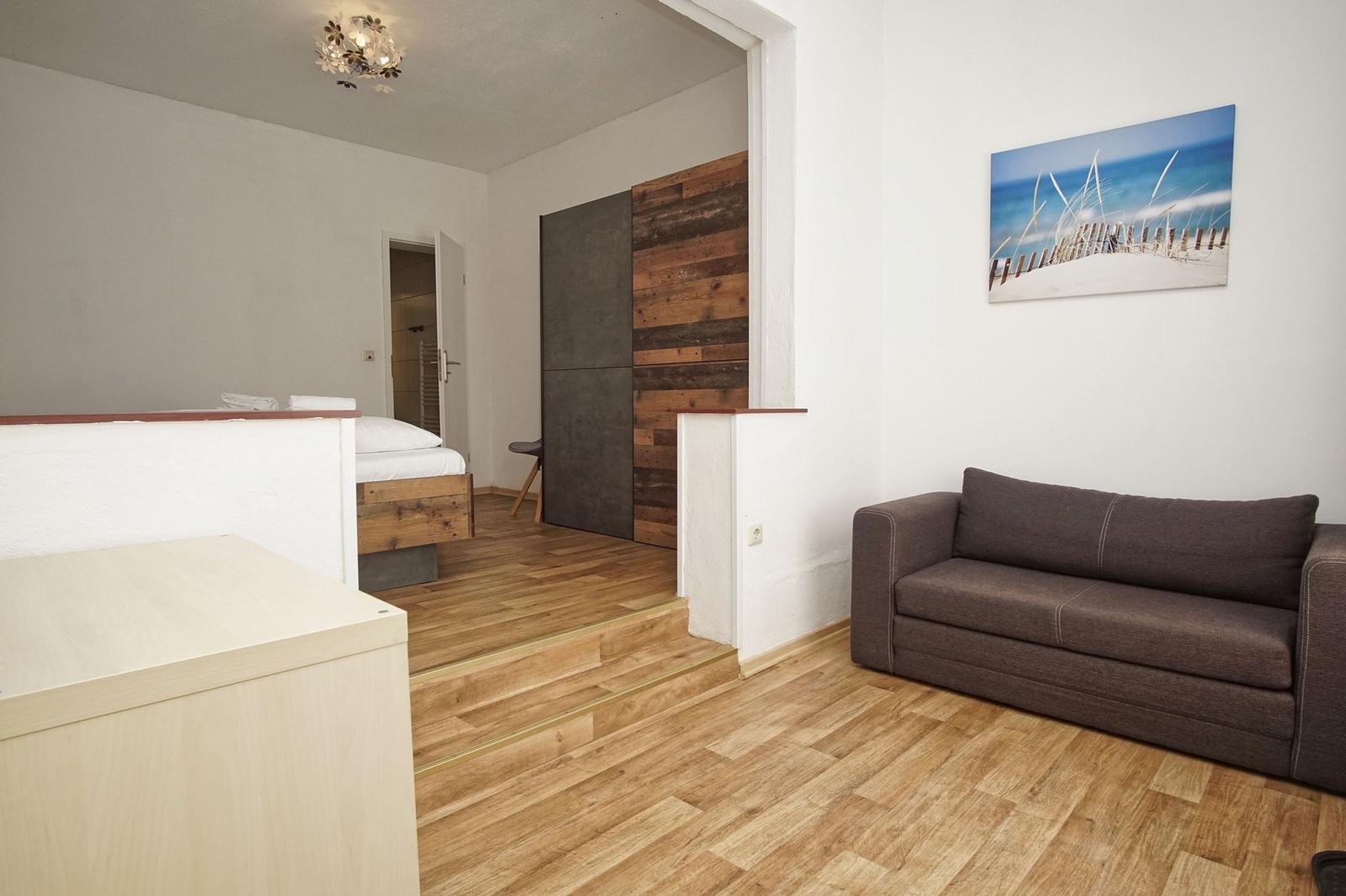 Wohnbereich mit Sofa und Blick in Schlafzimmer mit Bett und Holzwand.