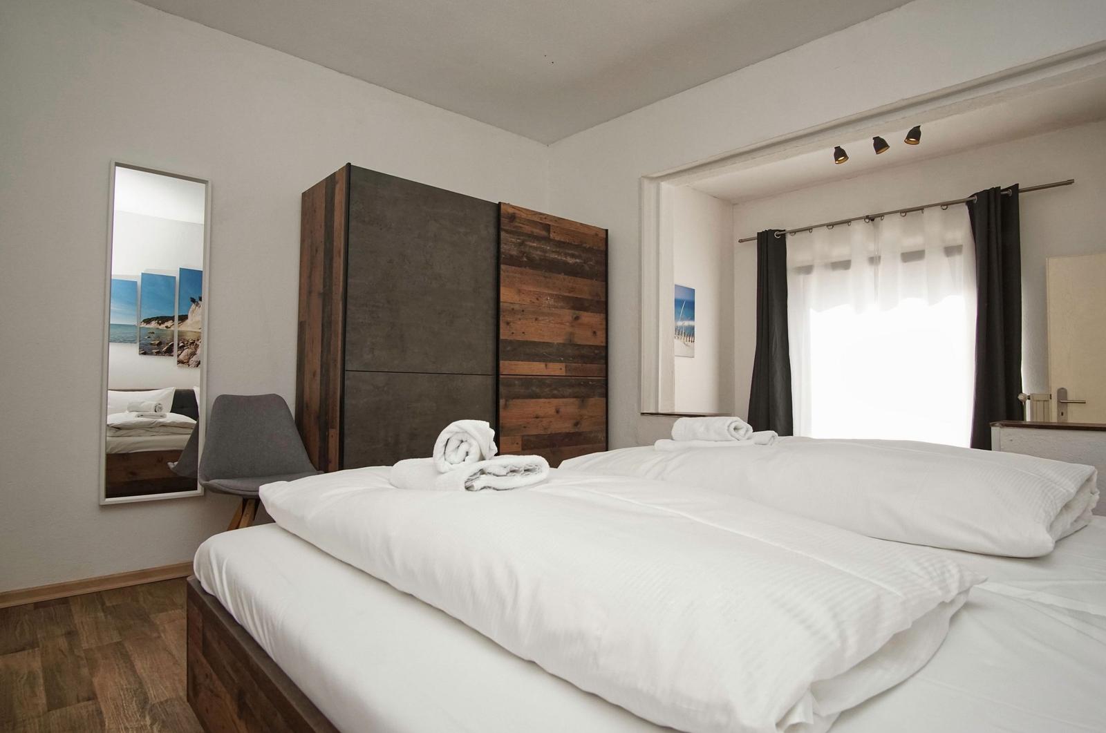 Schlafzimmer mit Doppelbett, Kleiderschrank und Spiegel. Fenster mit Vorhängen.