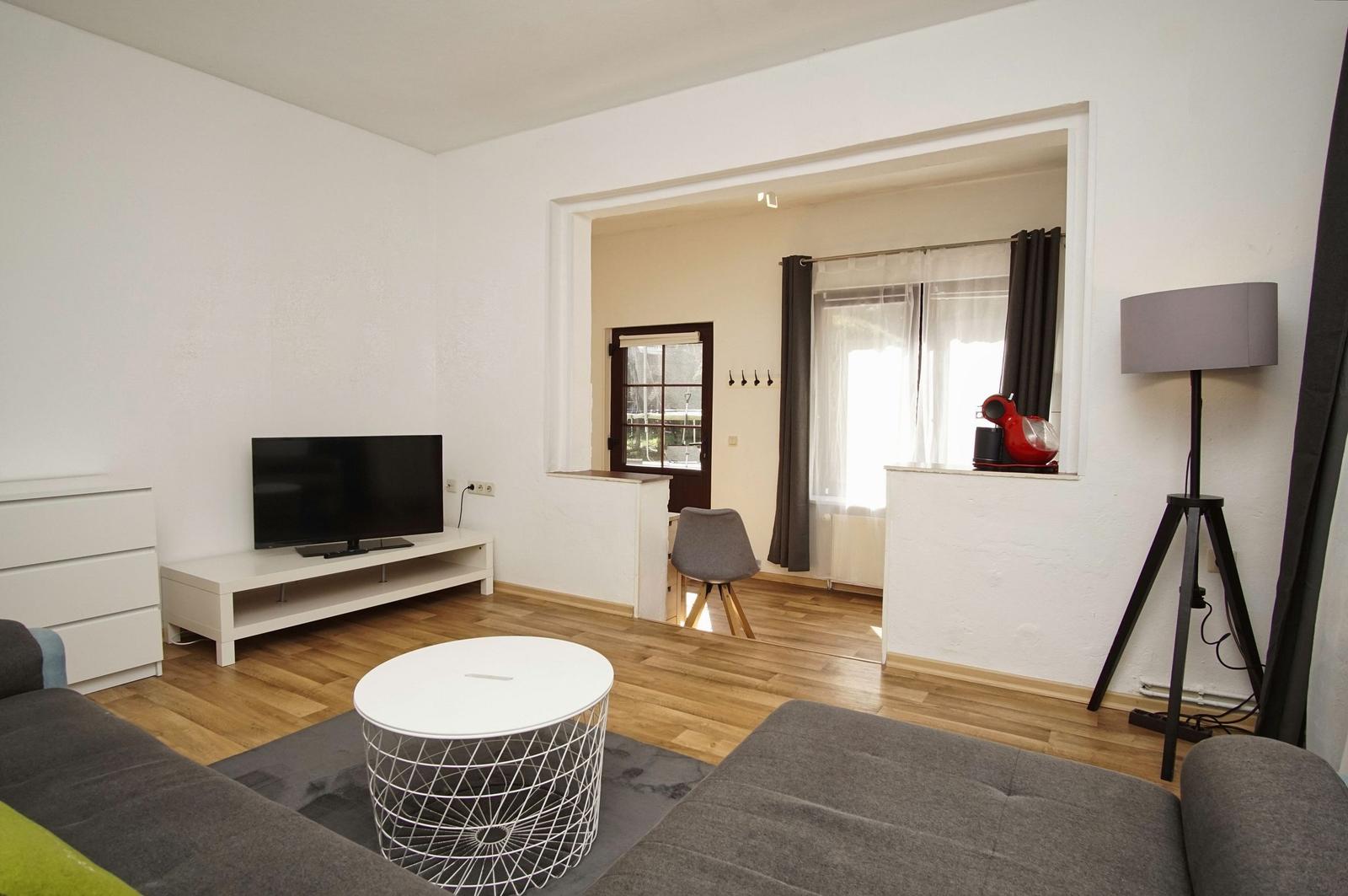 Wohnzimmer mit Sofa, TV und runden Tisch. Durchgang zu Küche mit Fenster.