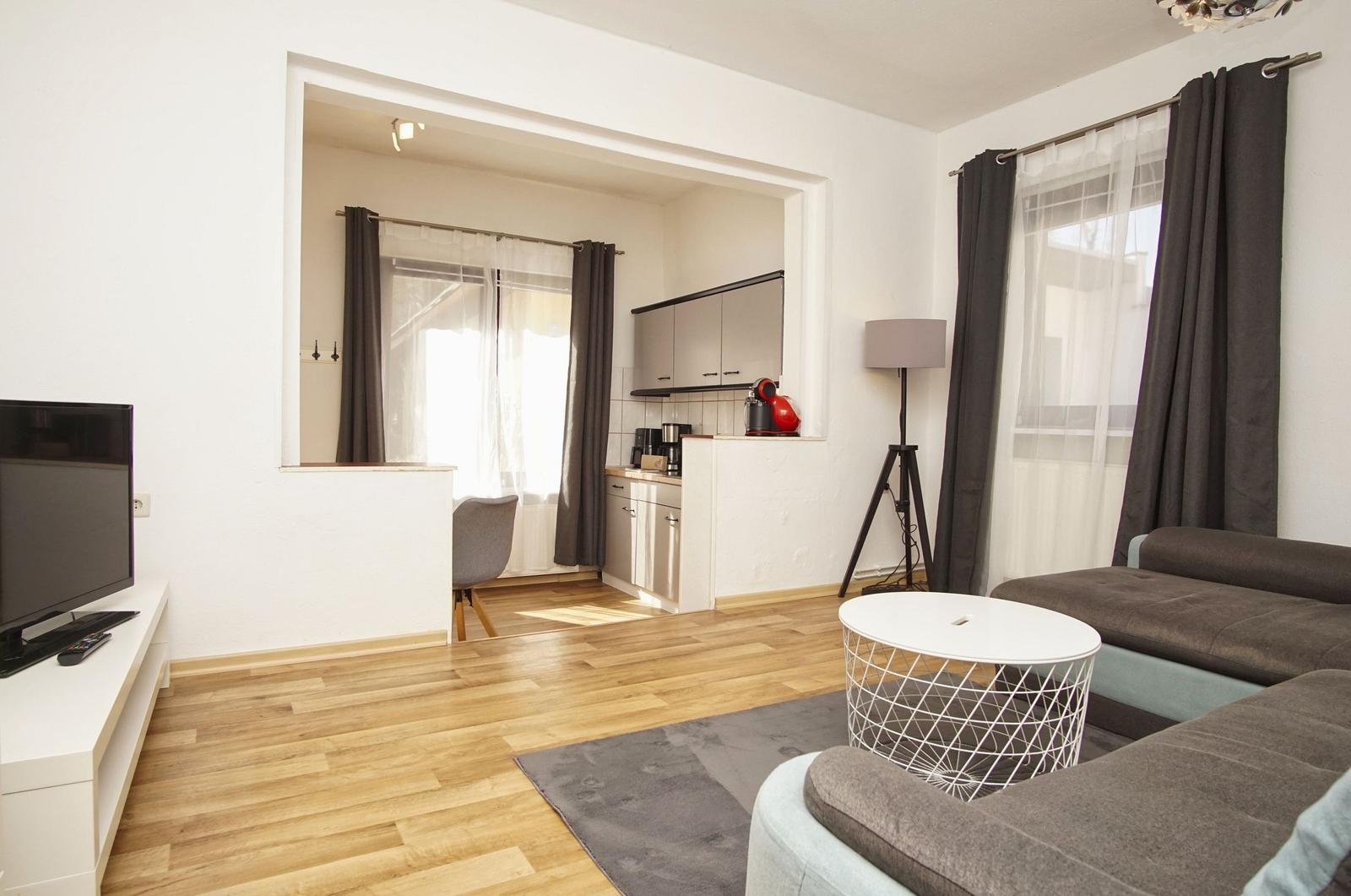 Wohnzimmer mit Sofa, Tisch und Fernseher. Küche im Hintergrund.