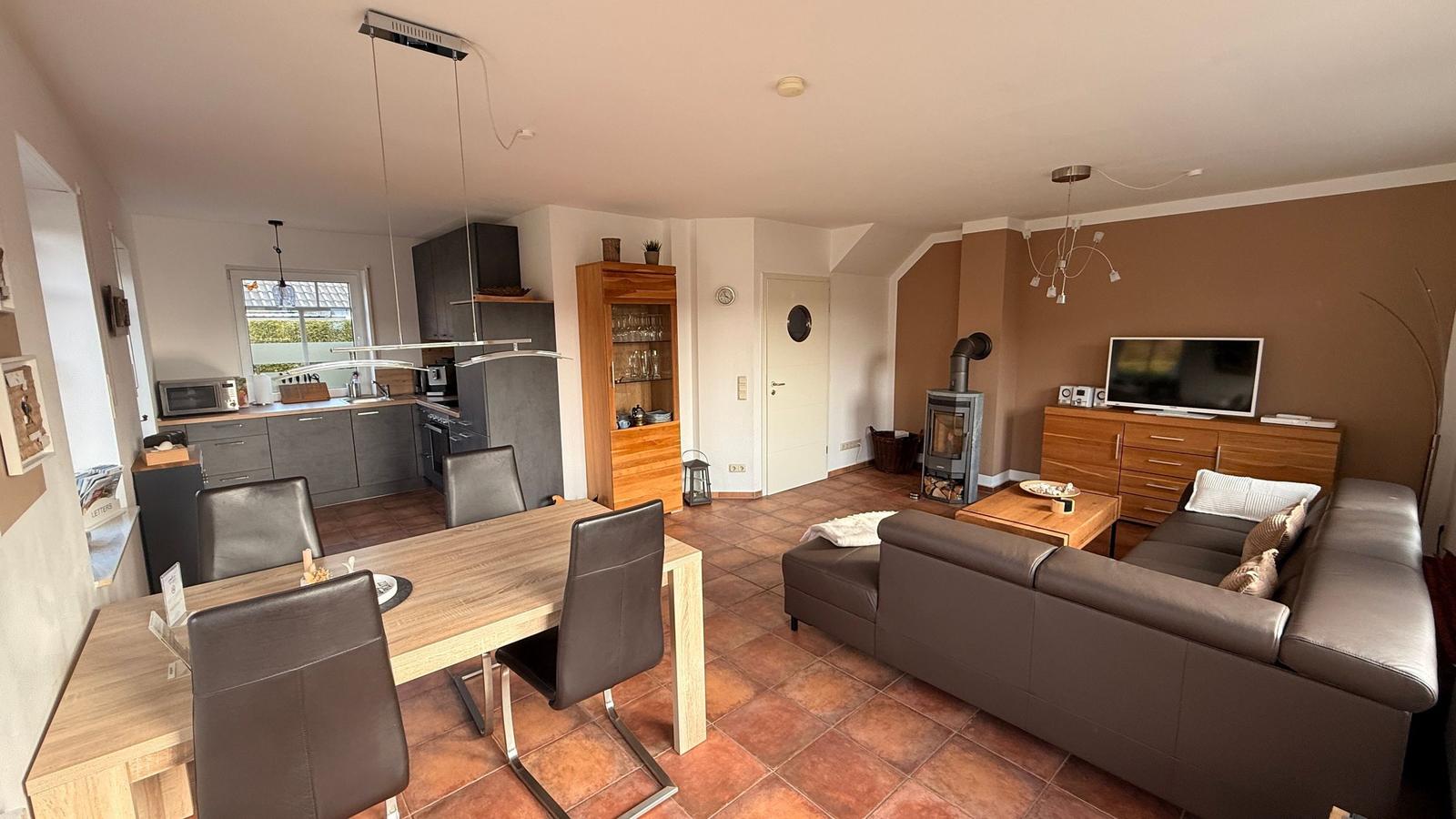 Wohn- und Essbereich mit Küche, Sofa und Holztisch. Terrasse und Kamin sichtbar.