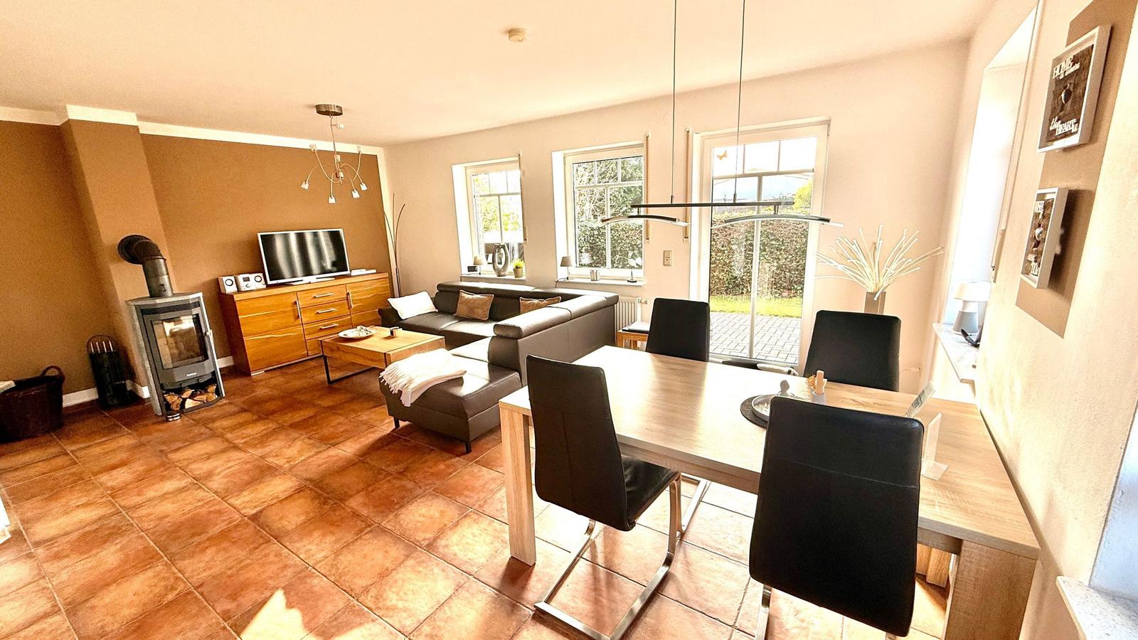 Wohn- und Essbereich mit Sofa, Esstisch und Kaminofen. Terrassentür führt nach draußen.