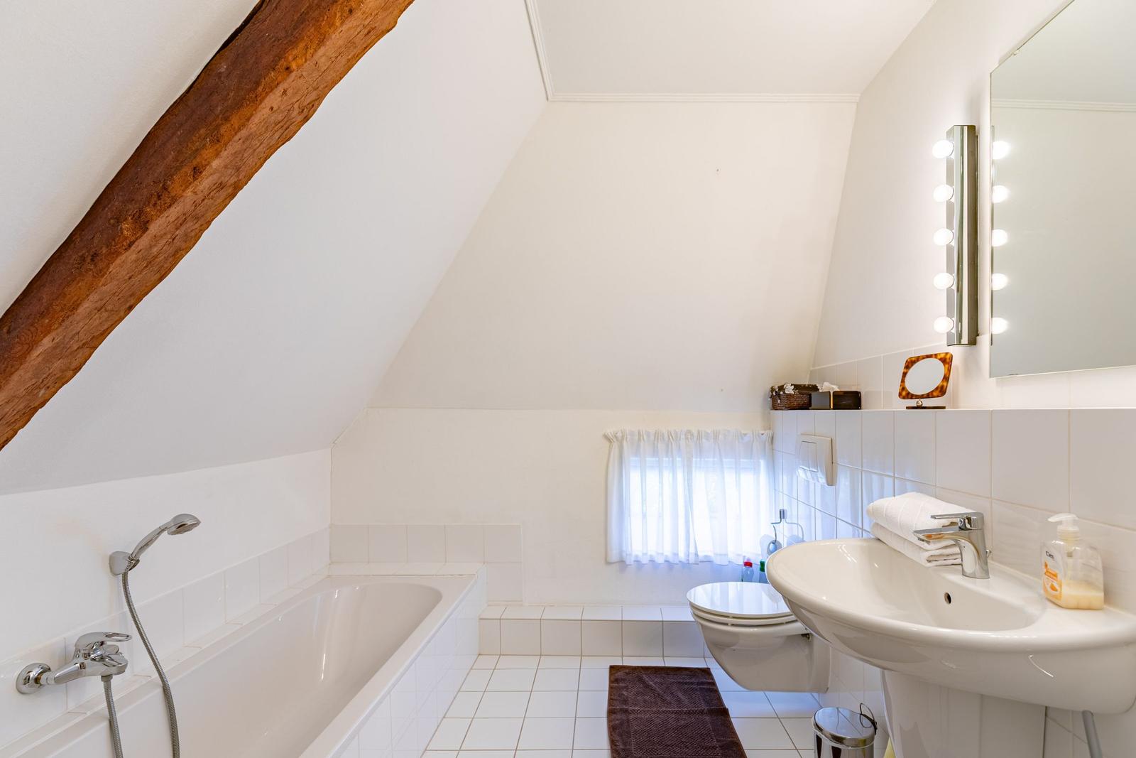 Badezimmer mit Badewanne, Toilette und Waschbecken unter schräger Decke mit Holzbalken.