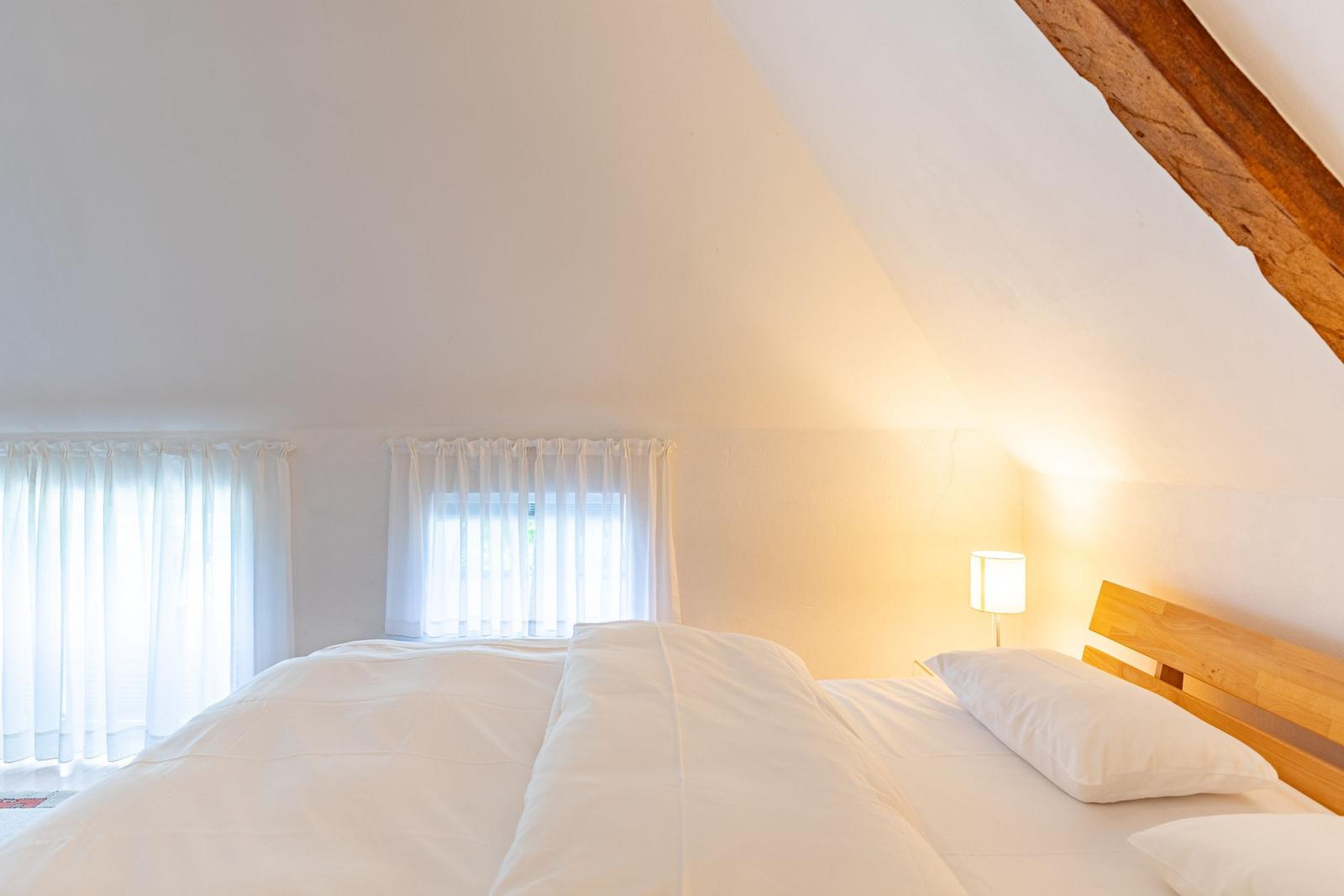 Schlafzimmer mit Dachschrägen, Bett mit weißen Laken und Holzkopfteil, zwei Fenster mit weißen Vorhängen.