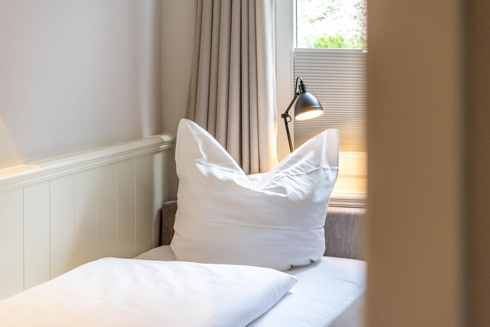 Ein Bett mit weißer Bettwäsche und Kissen. Eine Lampe und ein Fenster mit Jalousie.