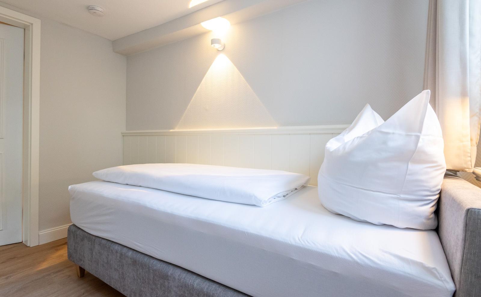 Ein Bett mit weißer Bettwäsche und Kissen. Wandbeleuchtung und Holzboden.
