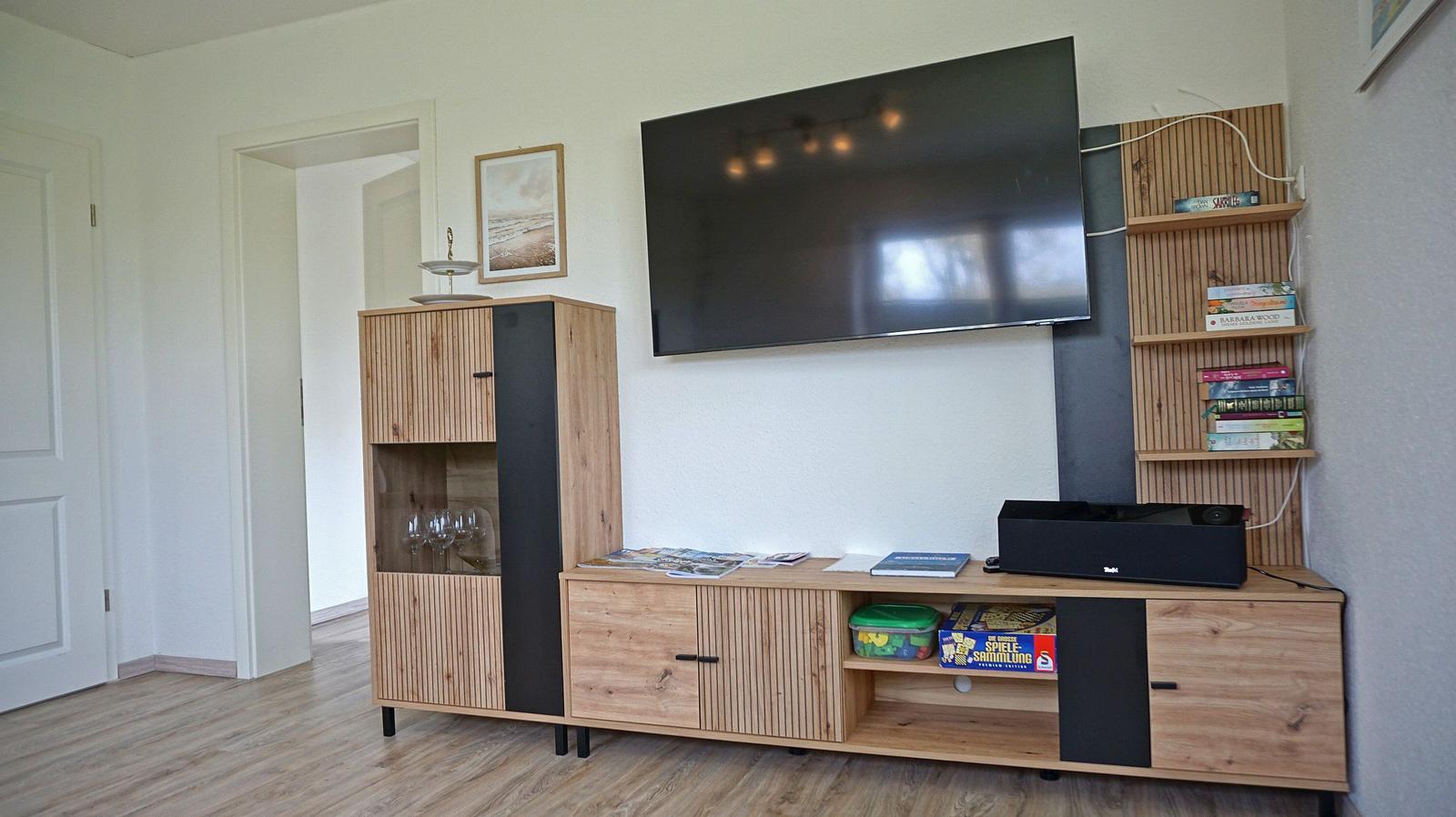 Wohnzimmer mit TV, Holzmöbel und Regalen. Flur und Tür sind sichtbar.