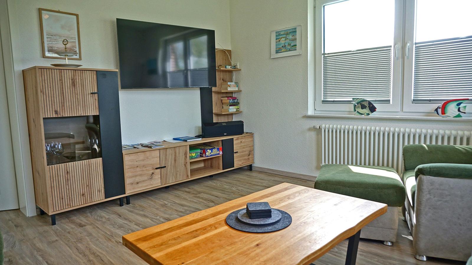 Wohnzimmer mit TV, Holzmöbel und grünem Sofa. Fenster mit Jalousien und Wandbildern.