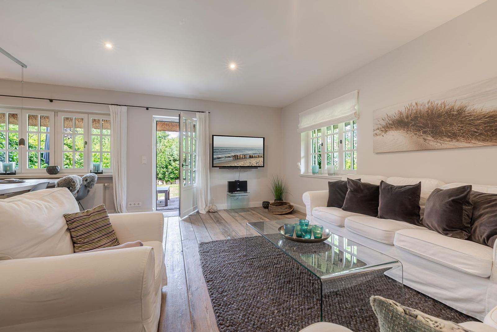 Wohnzimmer mit weißen Sofas, Glas-Tisch und Holzboden. Fernseher an der Wand. Tür führt nach draußen.