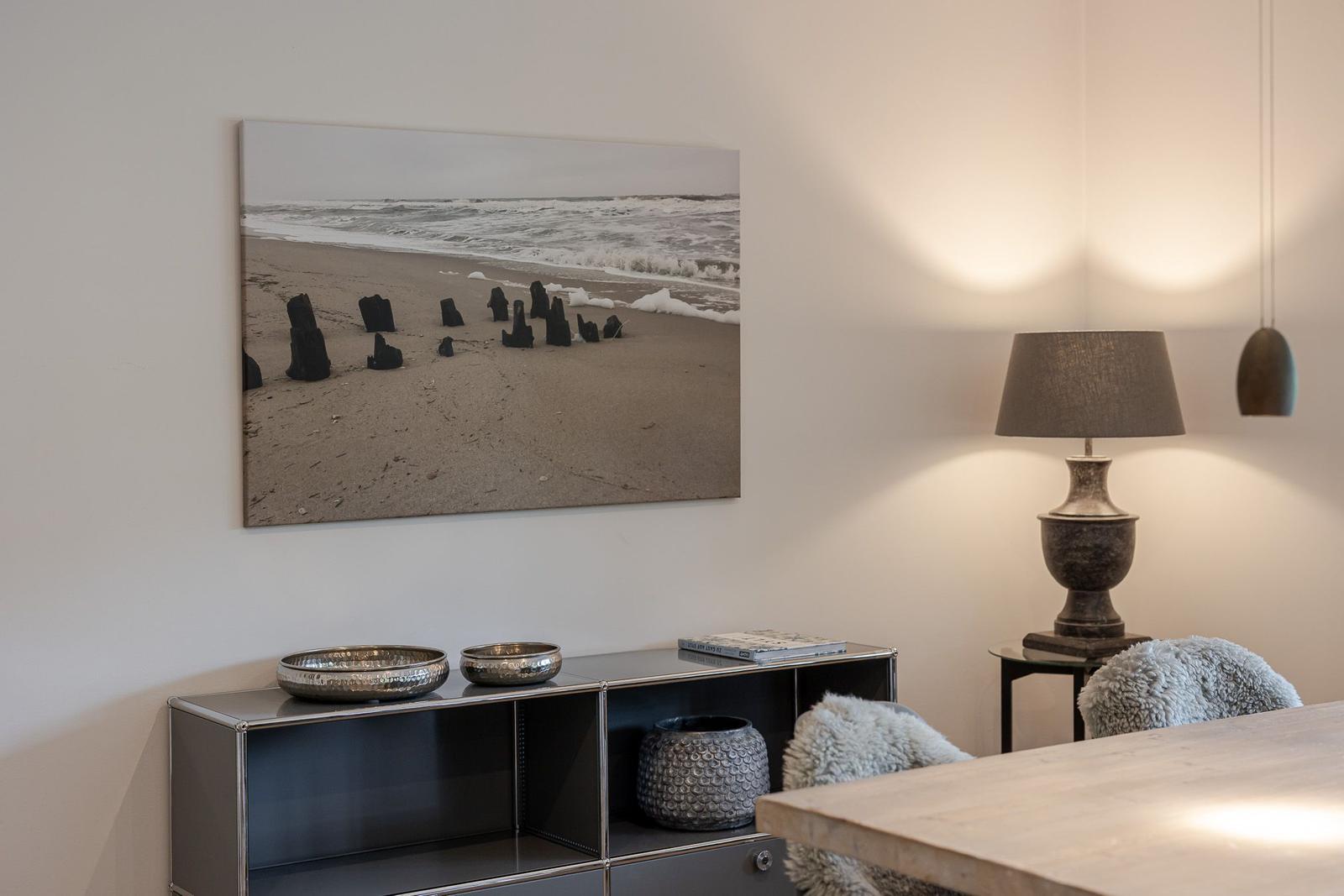 Wandbild zeigt Strand mit Pfählen, Tischlampe und Metallregal mit Schalen.