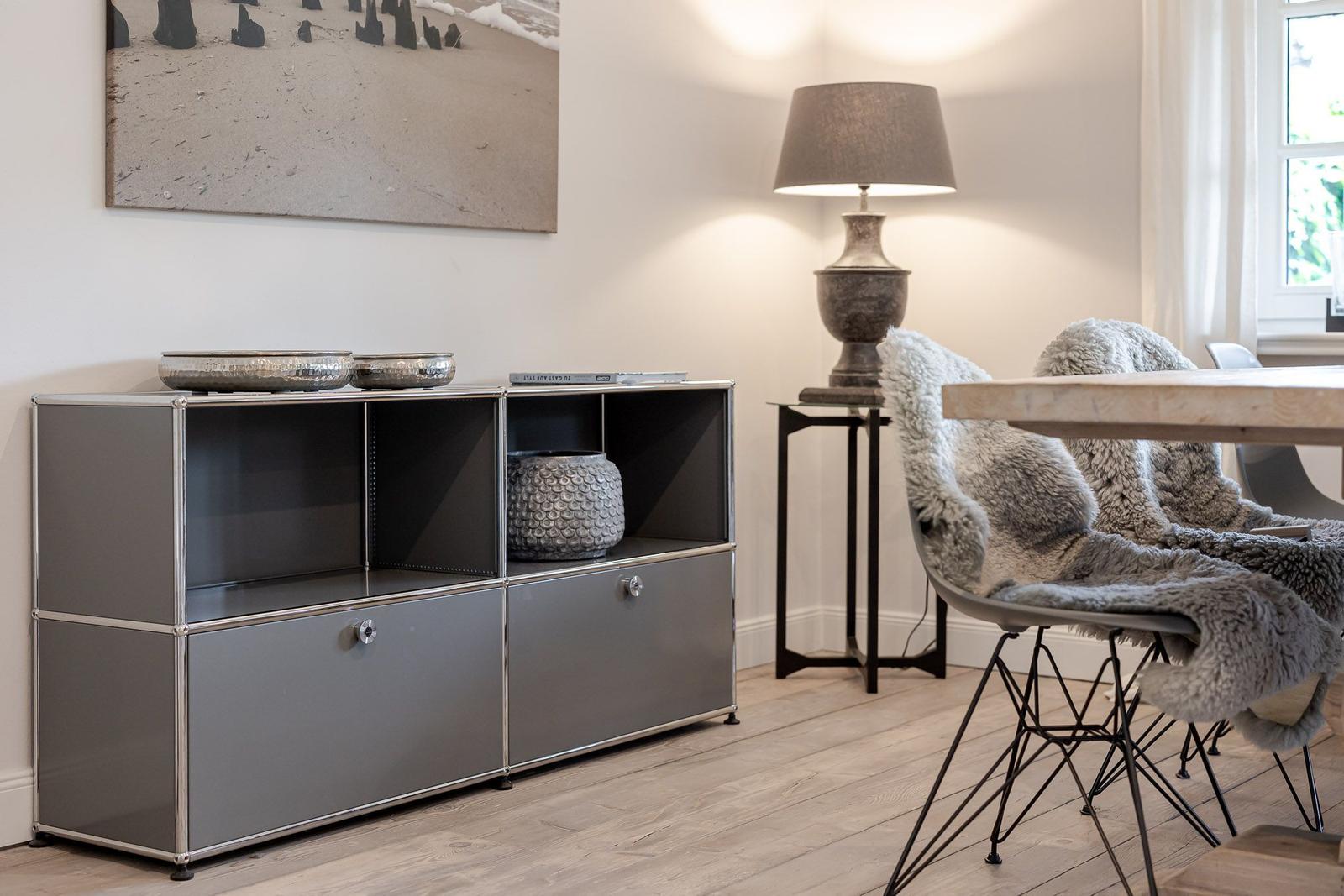 Ein Raum mit grauem Sideboard, Lampe und Stühlen mit Fellbezügen an einem Tisch.
