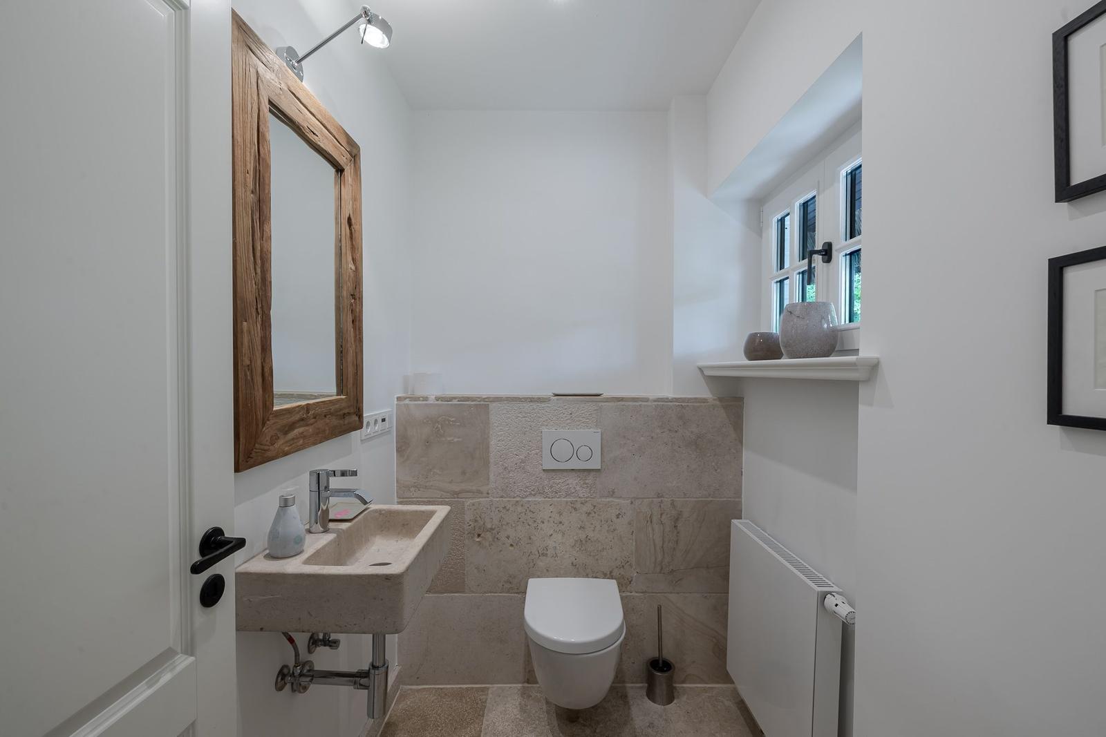 Badezimmer mit Waschbecken, Toilette und Holzspiegel. Steinwand und Fenster.