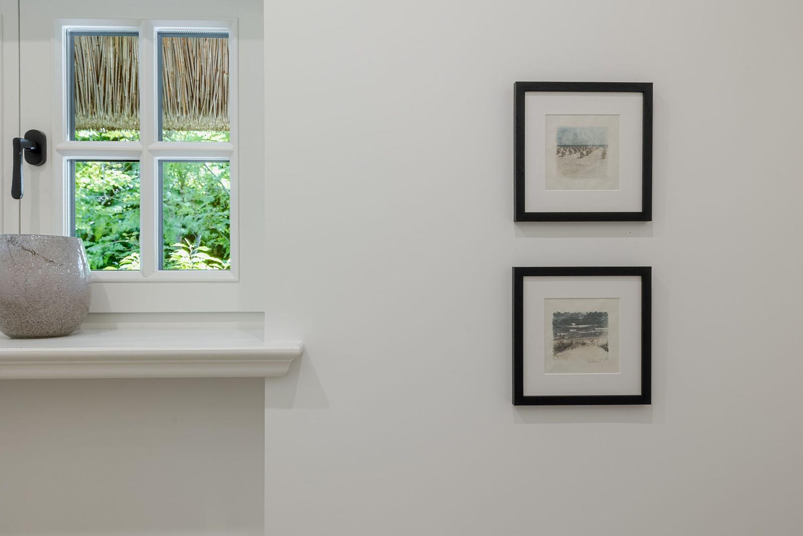 Fenster mit Blick auf grüne Pflanzen, Wand mit zwei gerahmten Bildern und Vase auf Fensterbank.