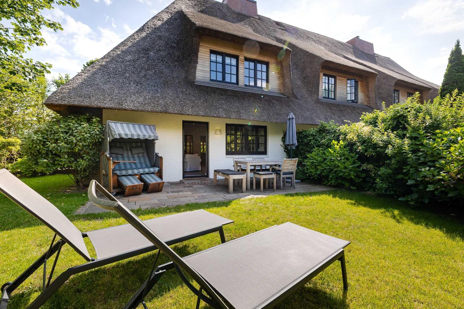 Haus mit Strohdach, Gartenbereich mit Liegestühlen und Terrasse mit Tisch und Stühlen.