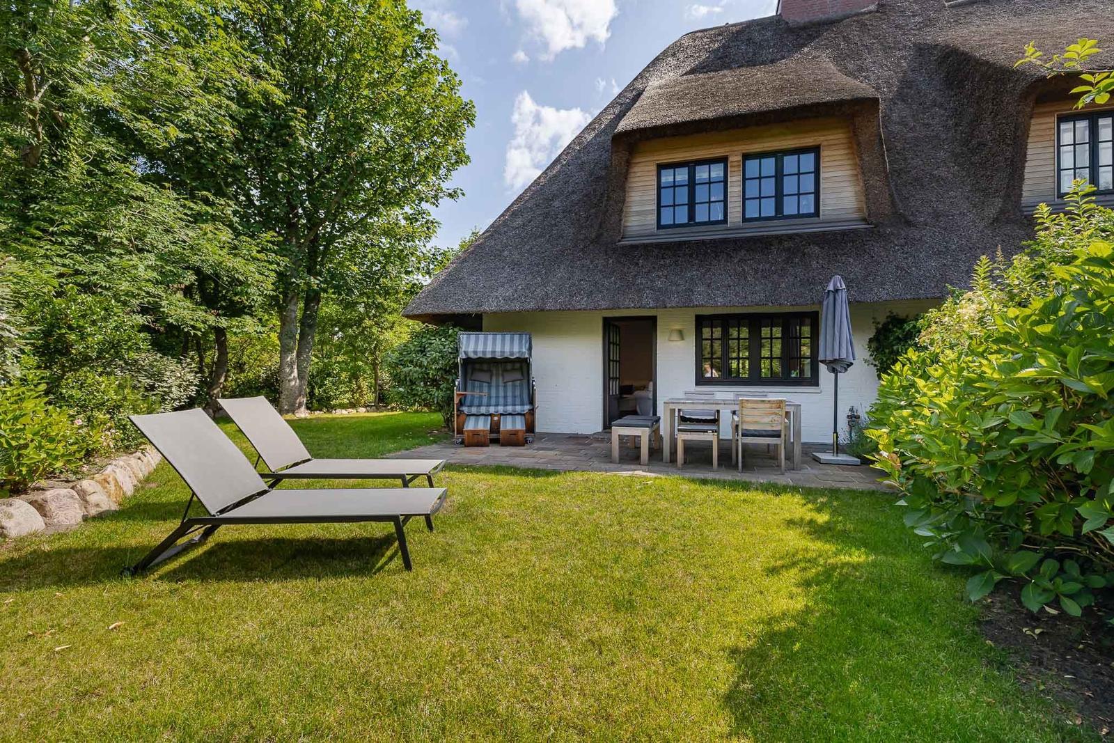 Haus mit Strohdach, Garten mit Liegestühlen und Terrasse mit Tisch und Stühlen.