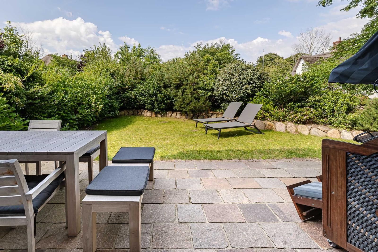 Hinterhof mit Terrasse, Tisch, Stühlen und Liegen. Grünflächen und Hecke im Hintergrund.