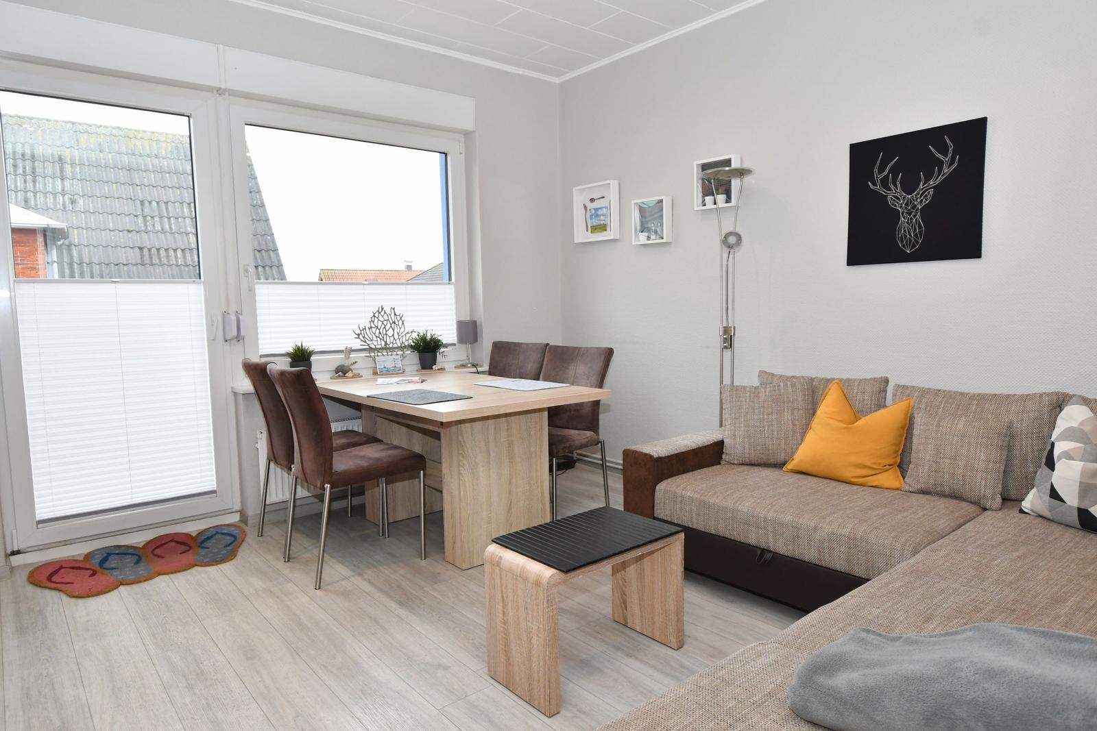Wohn- und Essbereich mit Sofa, Tisch und Stühlen. Fenster mit Jalousien und Wandbild.