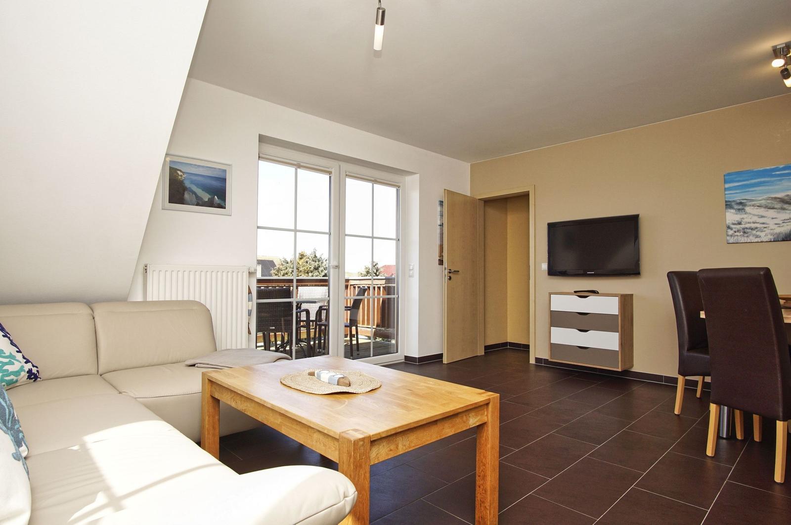 Wohnzimmer mit Sofa, Esstisch und TV. Balkon über Glastür. Holztisch und -stühle.