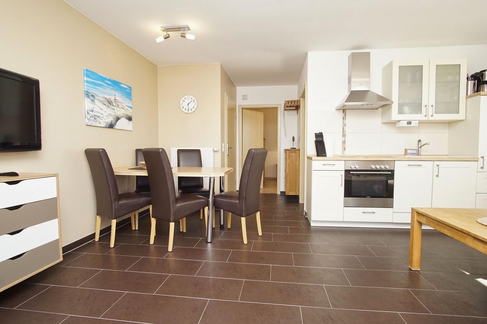 Küche mit Essbereich und Wohnzimmer. Weiße Schränke, Holztisch, Fernseher und Wanduhr.