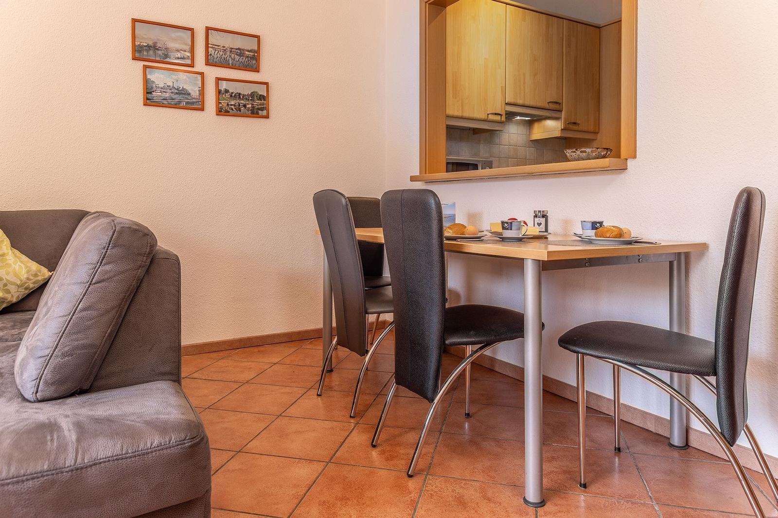 Essbereich mit Tisch, Stühlen und integrierter Küchenzeile. Sofa und Wandbilder sind sichtbar.