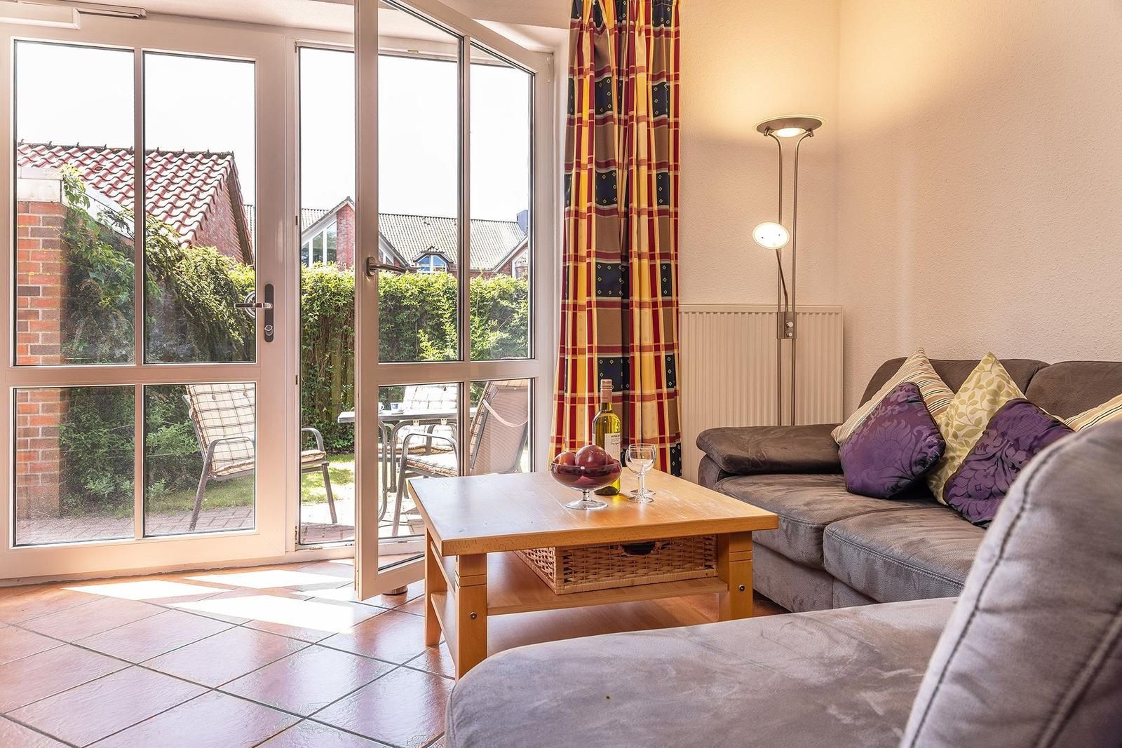 Wohnzimmer mit Sofa, Couchtisch und Terrassentür. Draußen befindet sich eine Terrasse mit Gartenmöbeln.