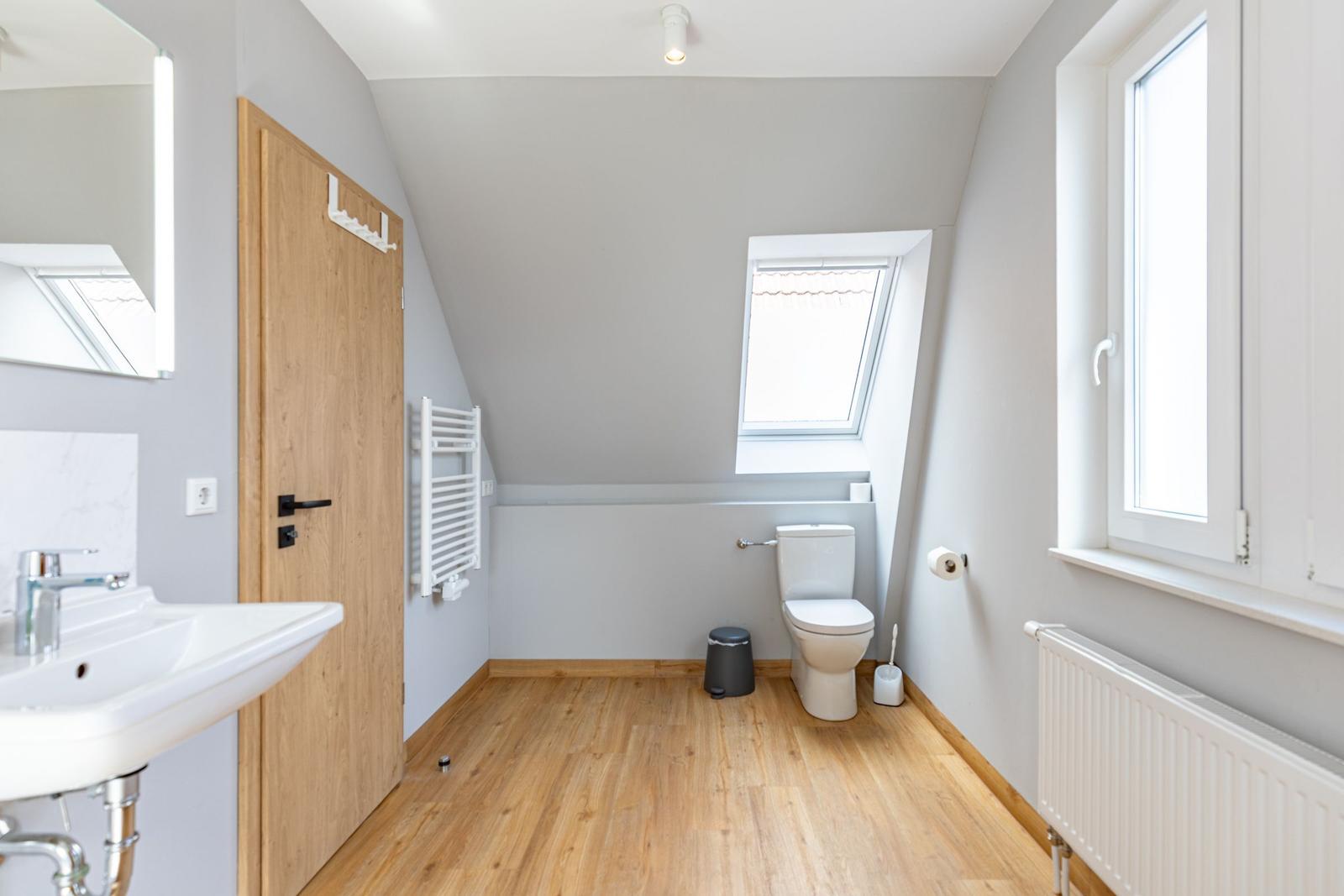 Bathroom with toilet, sink, and wooden floor. Windows provide natural light.
