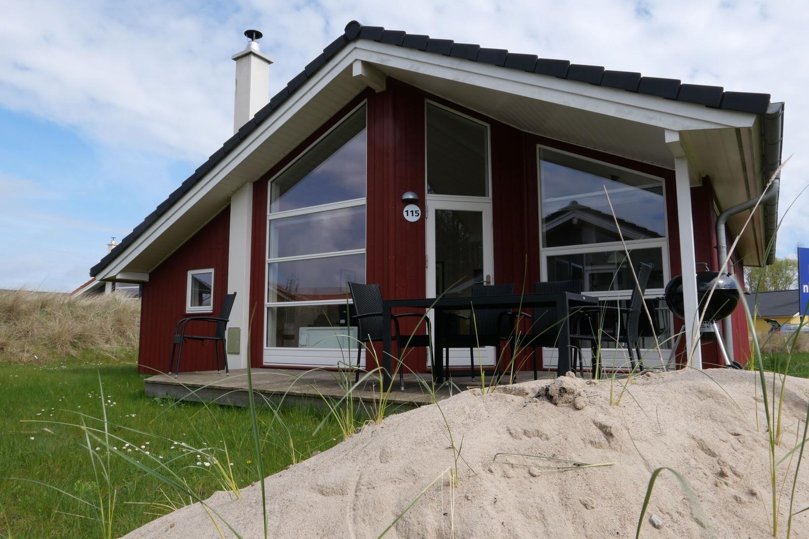 Rotes Ferienhaus mit Terrasse, Grill und großen Fenstern. Sandhügel im Vordergrund.