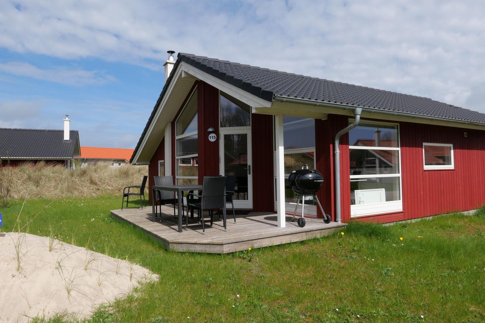 Rotes Ferienhaus mit Terrasse, Grill und Sitzgelegenheiten im Gras.