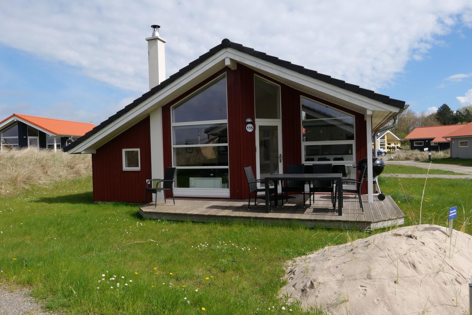 Rotes Ferienhaus mit Terrasse, Grill und großem Fenster. Grasland und Sandhügel im Vordergrund.