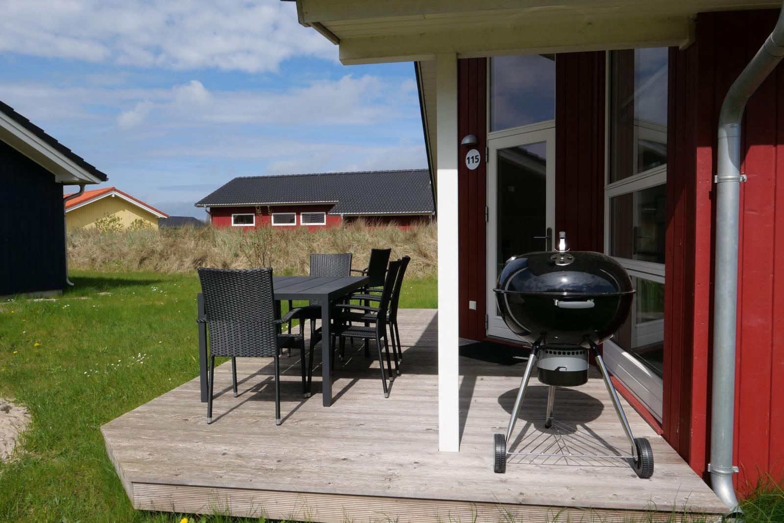 Terrasse mit Tisch, Stühlen und Grill vor rotem Haus mit Hausnummer 115.