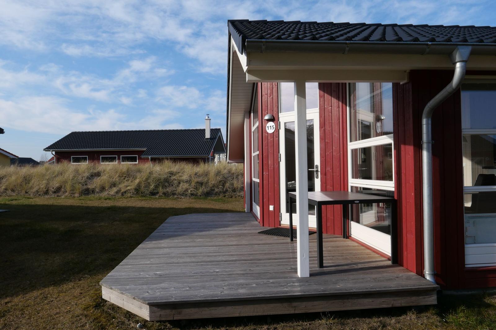 Rotes Ferienhaus mit Holzterrasse und Tisch. Hintergrund: Grasland und weiteres Haus.