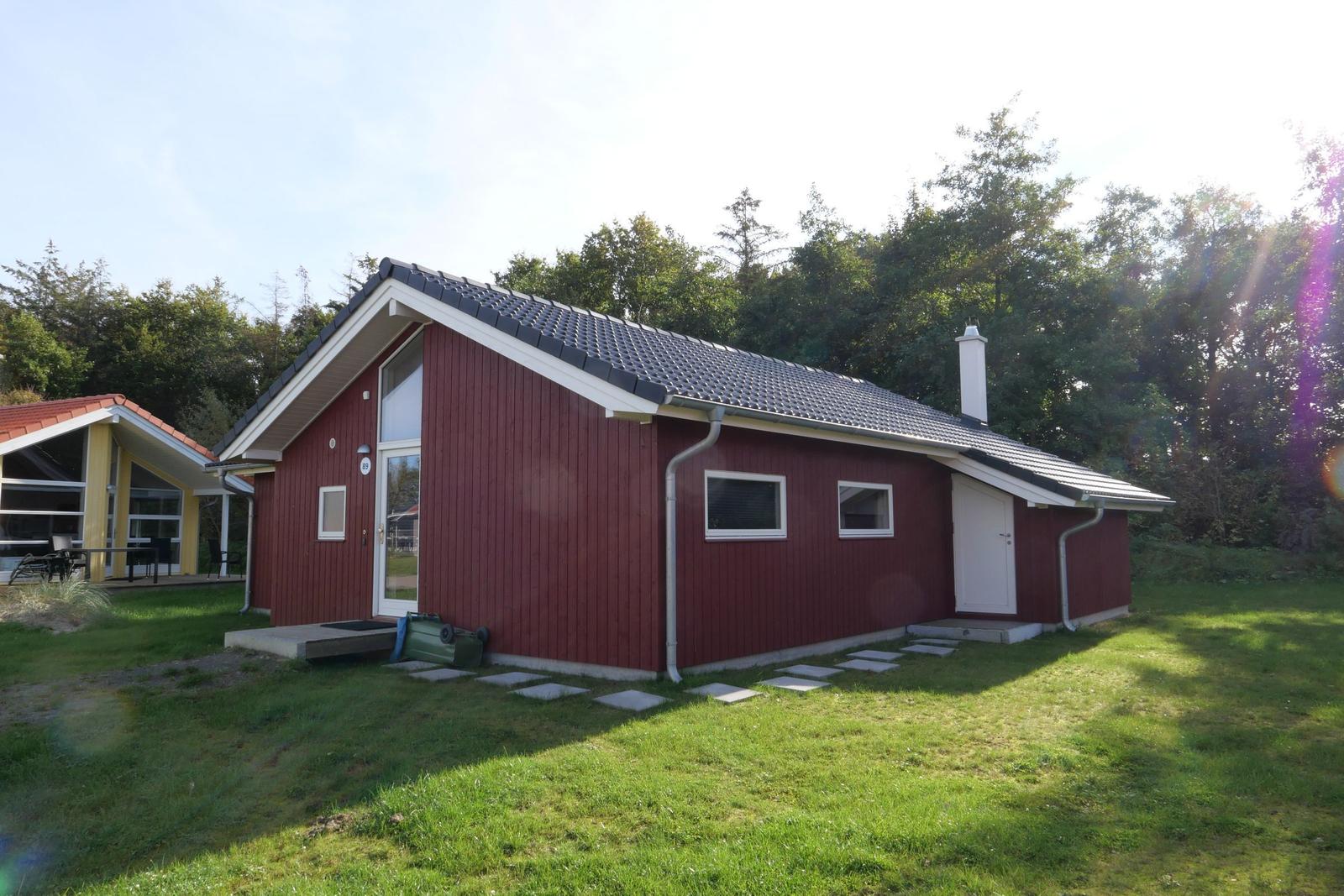 Rotes Haus mit grauem Dach, weißer Tür und Fenstern. Grasfläche und Bäume im Hintergrund.
