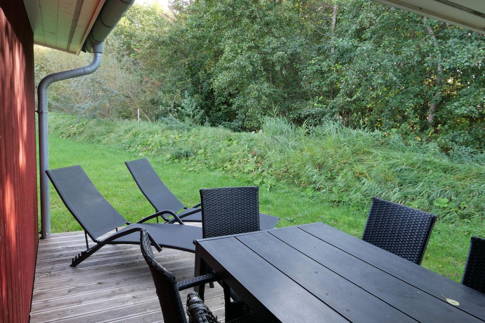 Terrasse mit Tisch, Stühlen und Liegestühlen. Hintergrund: Gras und Bäume.