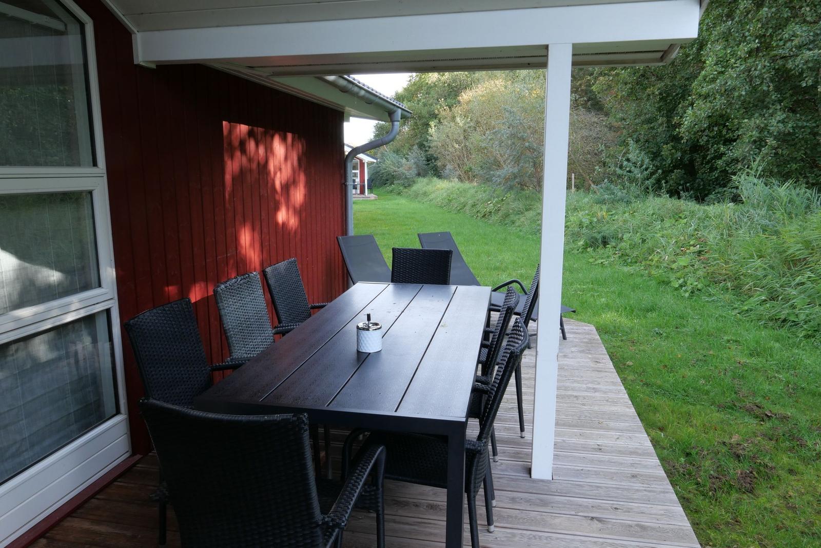 Terrasse mit Tisch und Stühlen neben rotem Haus mit Grünflächen im Hintergrund.