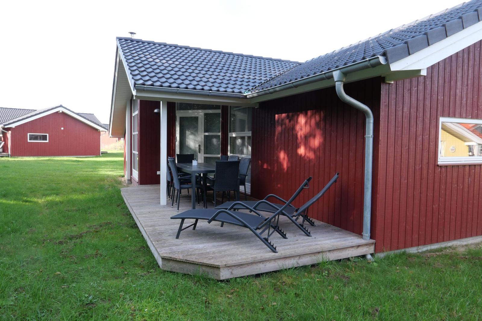 Roter Holzhaus mit Terrasse, Tisch, Stühlen und Liegestühlen. Grasfläche im Hintergrund.