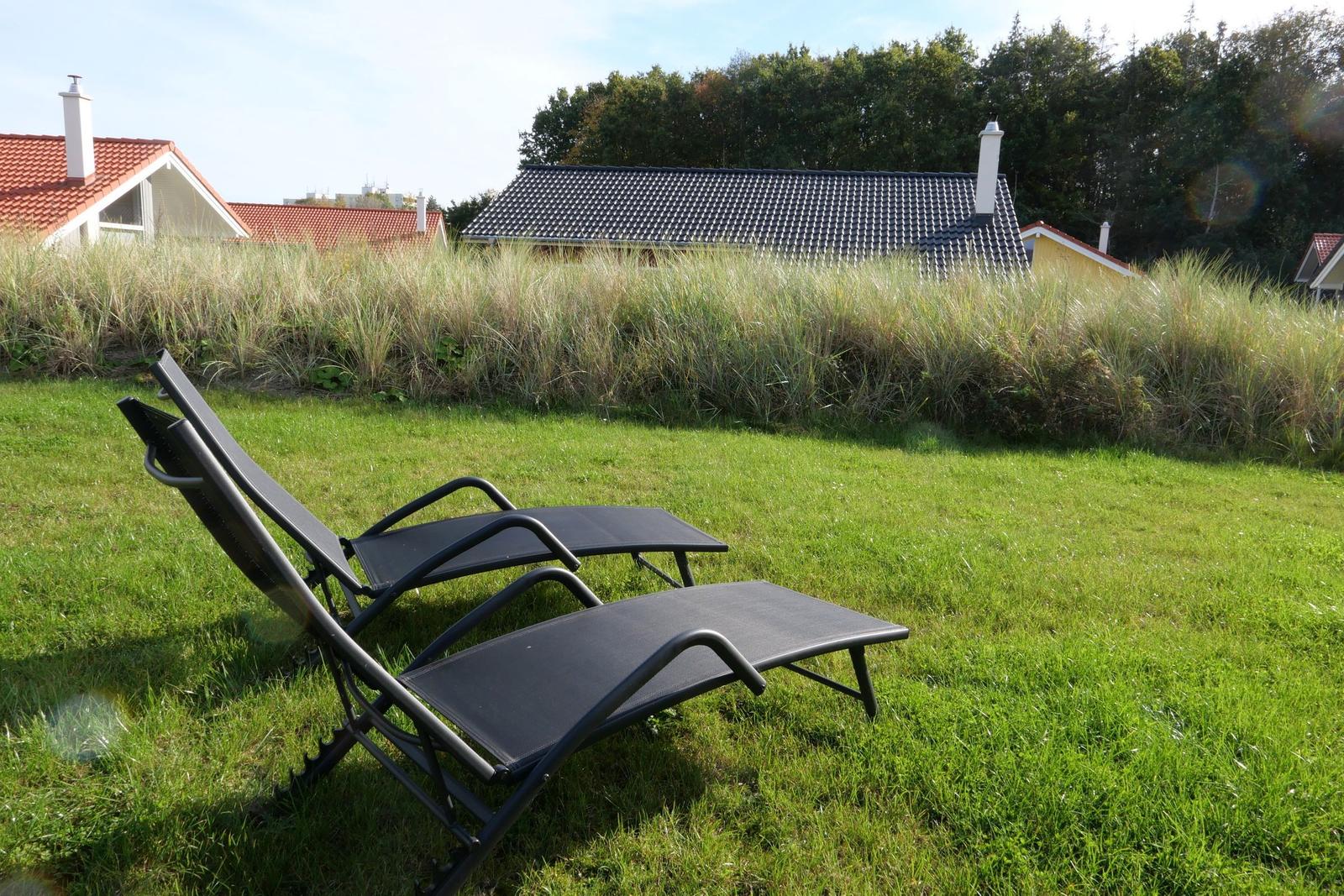 Two black lounge chairs on green lawn in front of hedges and houses.