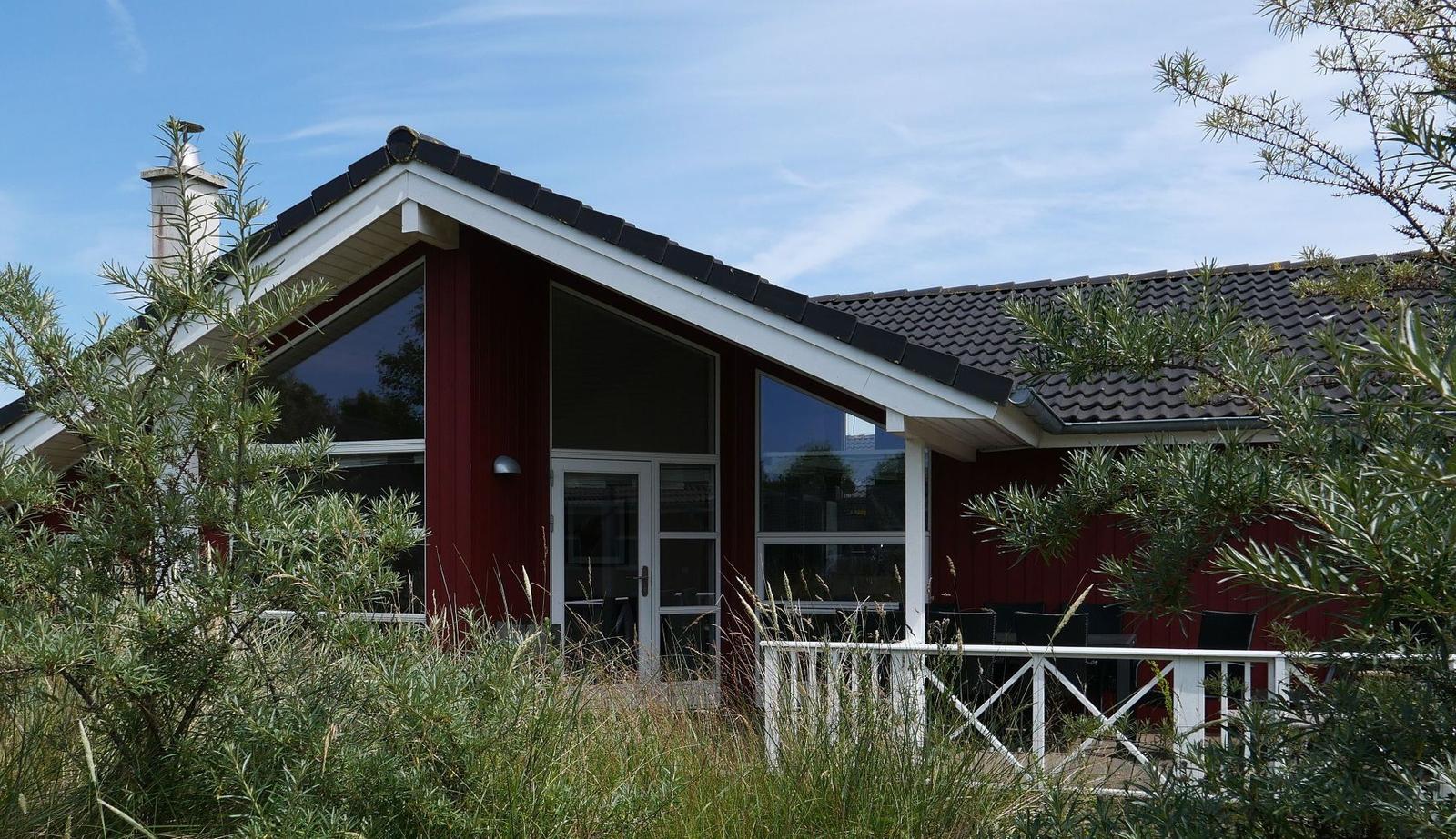 Rotes Haus mit weißem Geländer und großen Fenstern. Außenbereich mit Sitzgelegenheiten.