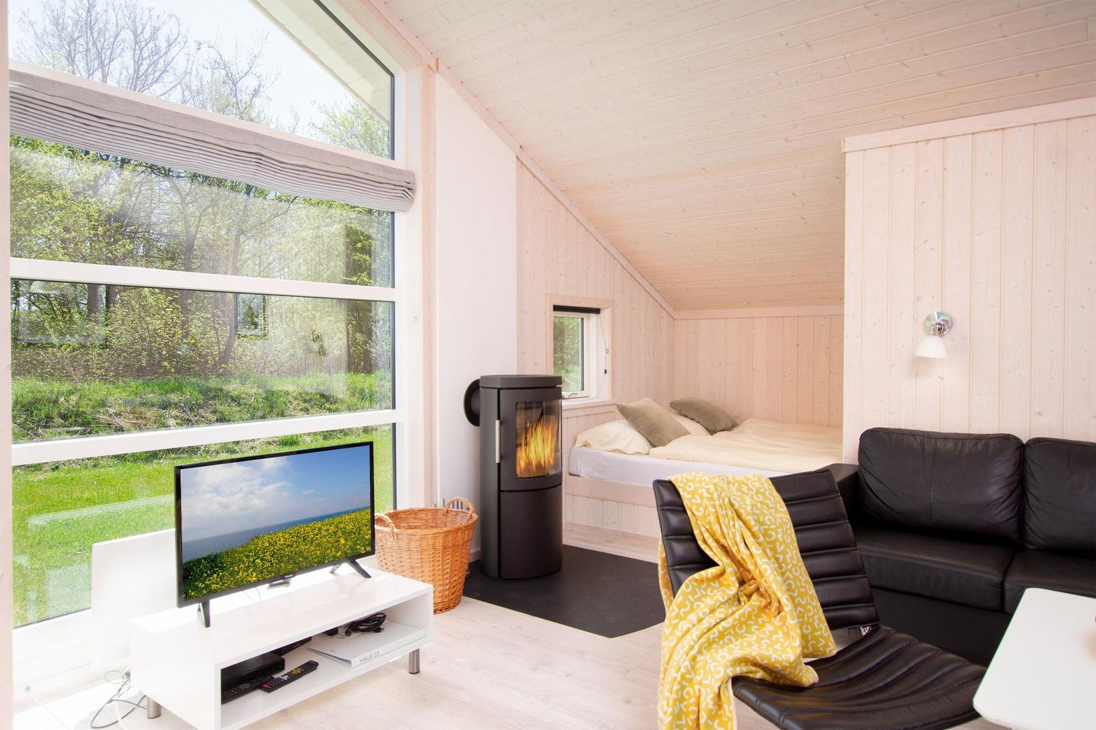 Ein Raum mit Sofa, Kamin und Bett unter Holzdecke. Große Fenster mit Blick auf Grün.