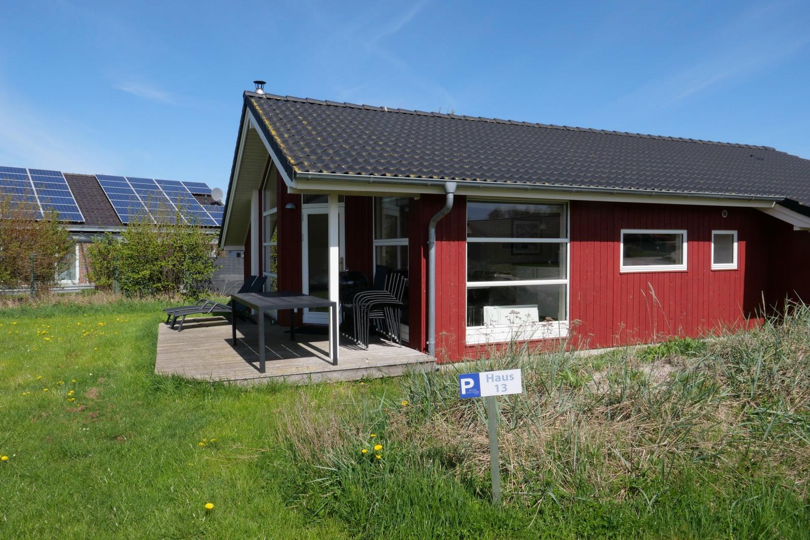 Rotes Haus mit Terrasse und Garten. Schild zeigt Haus 13. Hintergrund mit Solaranlagen.