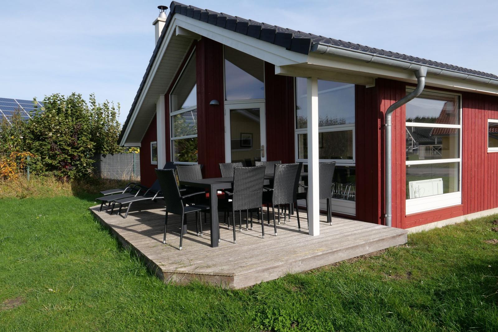 Rotes Haus mit Terrasse, Tisch, Stühlen und Liegestühlen. Grasfläche und Solaranlage im Hintergrund.