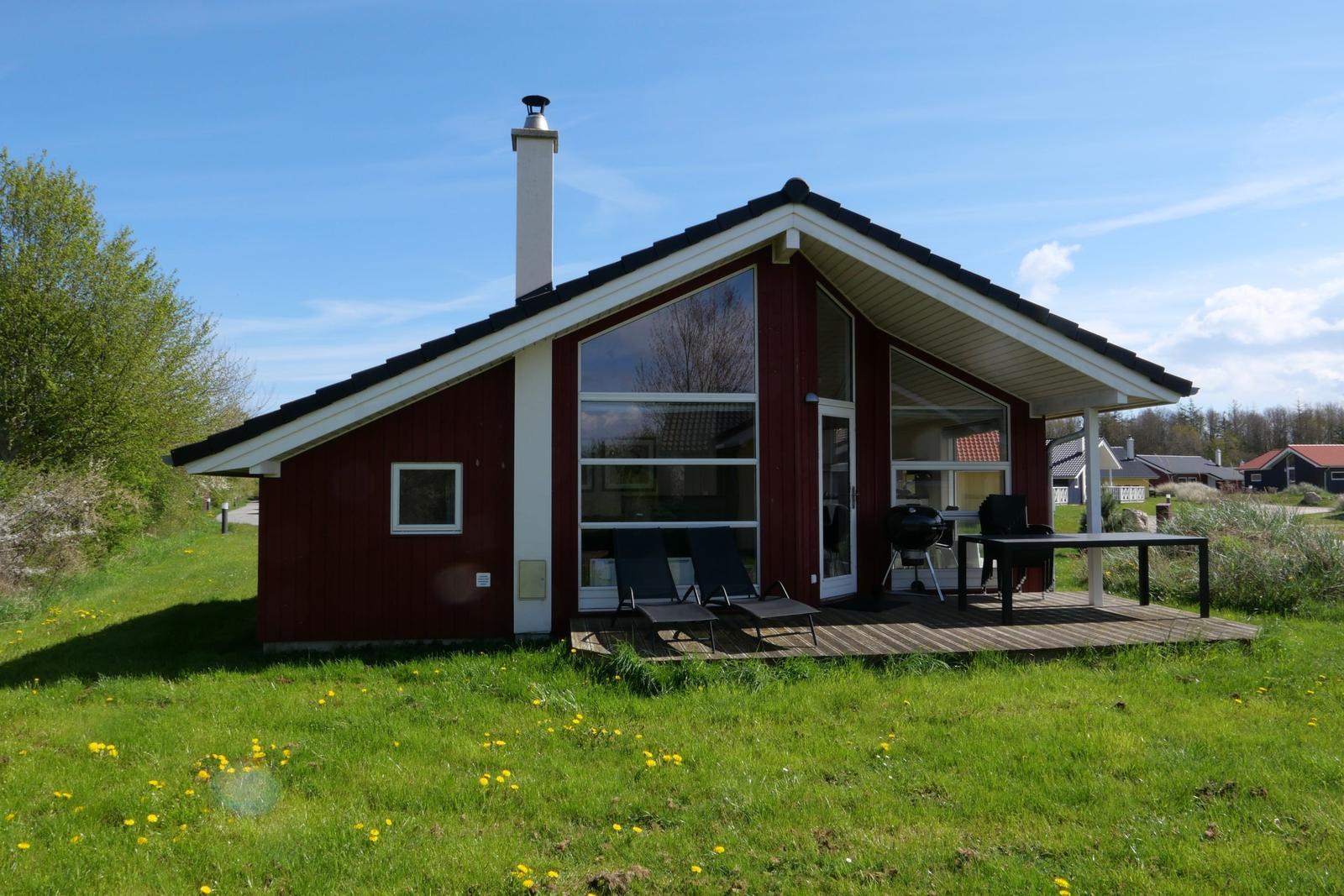 Rotes Haus mit Terrasse, Grill und Liegestühlen auf grünem Grund.