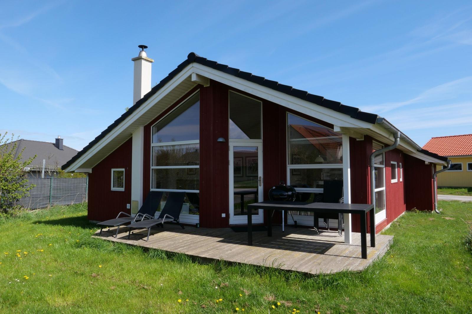 Rotes Haus mit Holzterrasse, Sonnenliegen und Grill. Große Fensterfront. Grün umgeben.