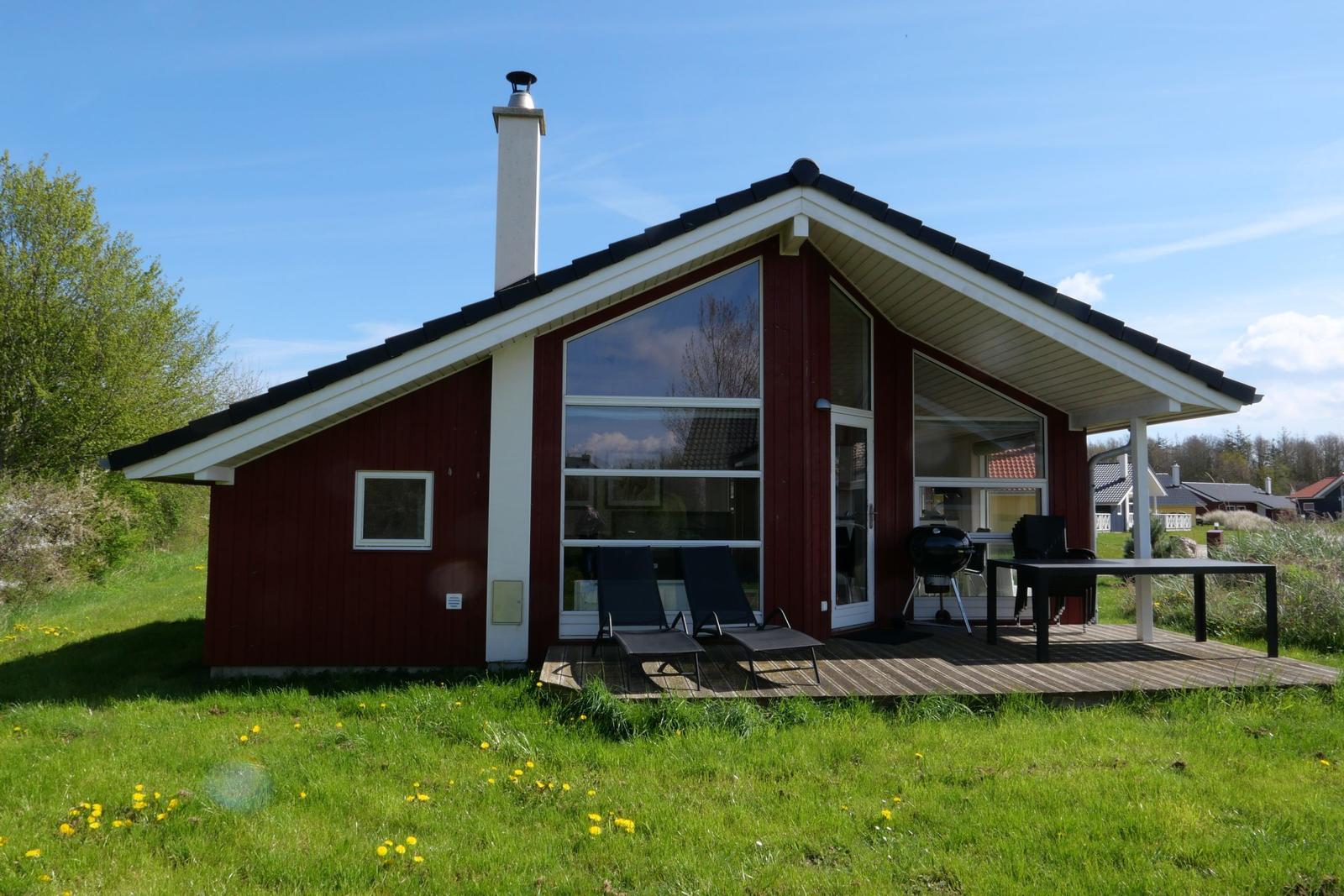 Rotes Ferienhaus mit Terrasse, Grill und Liegen. Grün umgeben, klarer Himmel.