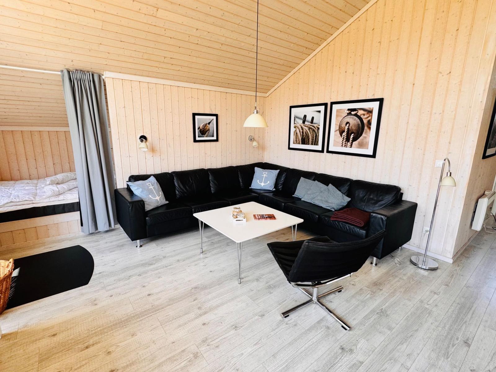 Wohnzimmer mit schwarzer Eckcouch, weißem Tisch und Holzwand. Schlafbereich im Hintergrund.